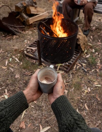 Two hands holding a mug of hot chocolate in front of a lit fire pit