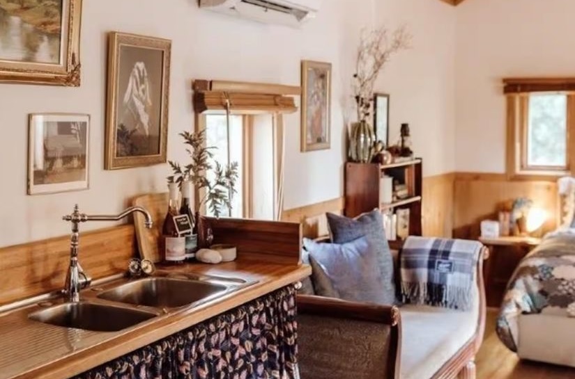 The cottage living space with a timber pitched ceiling, framed pictures on the walls, a leather armchair with a tartan blanket, and a kitchen sink in the foreground